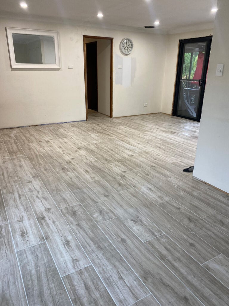 An interior view of a room undergoing renovation, featuring light grey wood-look floor tiles laid in a staggered plank pattern.