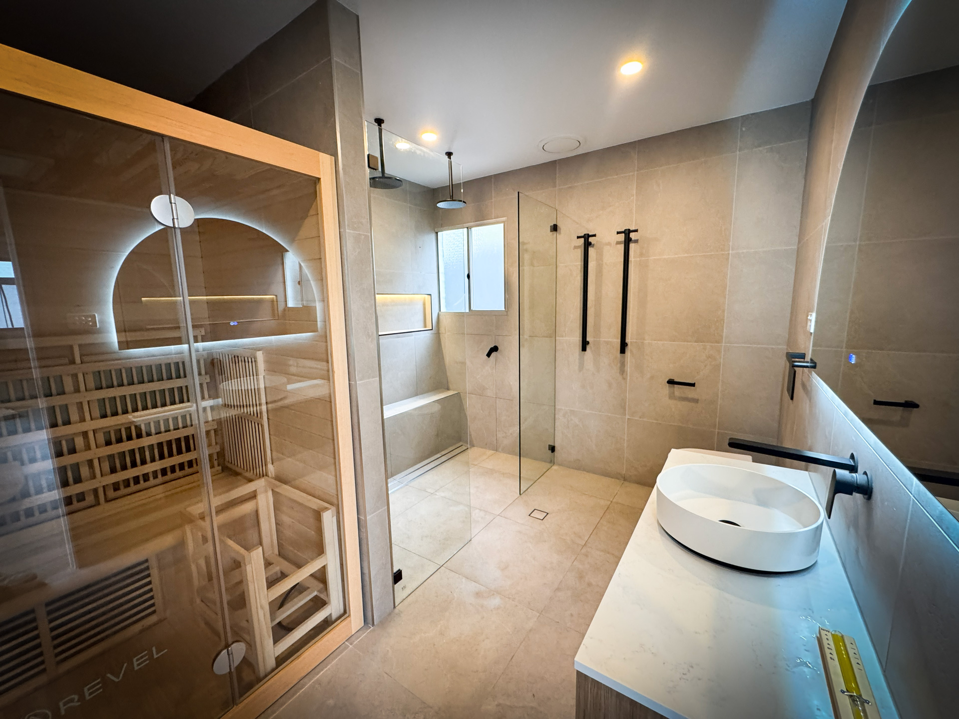 A modern bathroom featuring a wooden sauna cabin with a glass door, a large glass-enclosed walk-in shower, and a white vessel sink on a stone vanity.