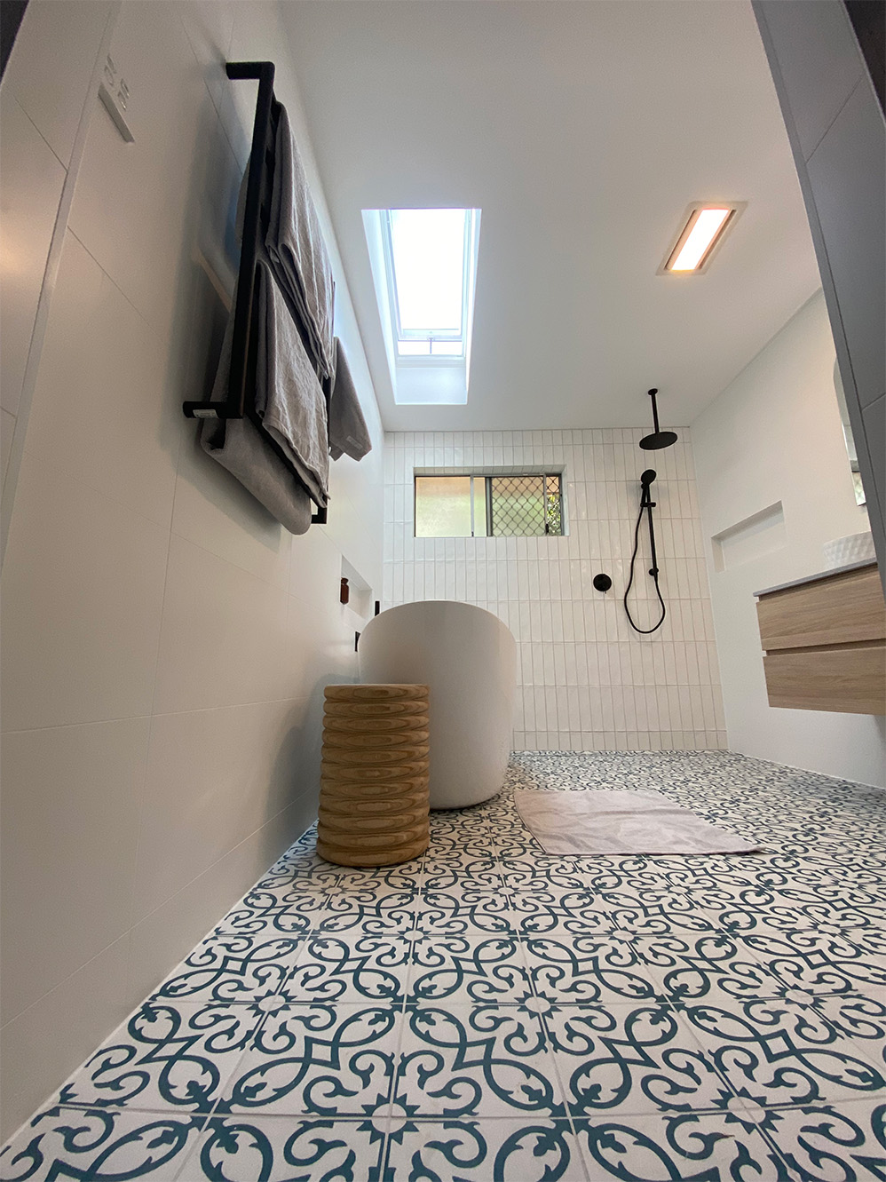 A low-angle view of a modern bathroom with patterned blue and white floor tiles, a white freestanding bathtub, and a wood-textured vanity.