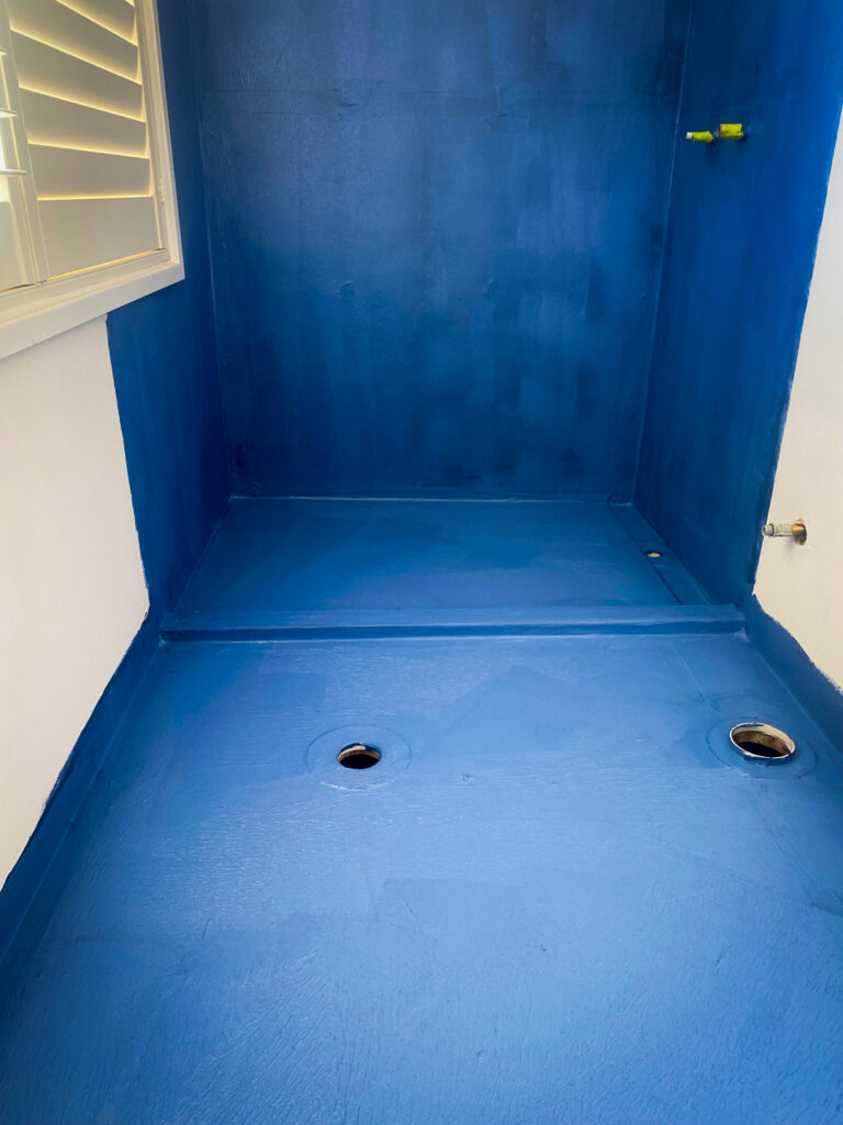 A bathroom shower area undergoing renovation, with the walls and floor coated in a vibrant blue liquid waterproofing membrane.