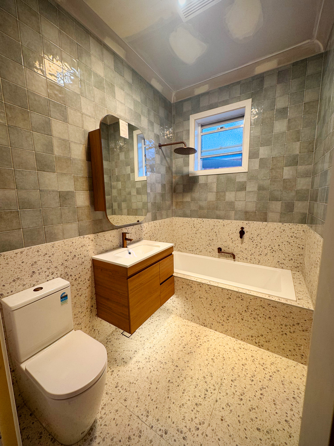 Modern bathroom featuring sage green square wall tiles, terrazzo floor and wainscoting, a floating wood vanity, and a built-in bathtub.