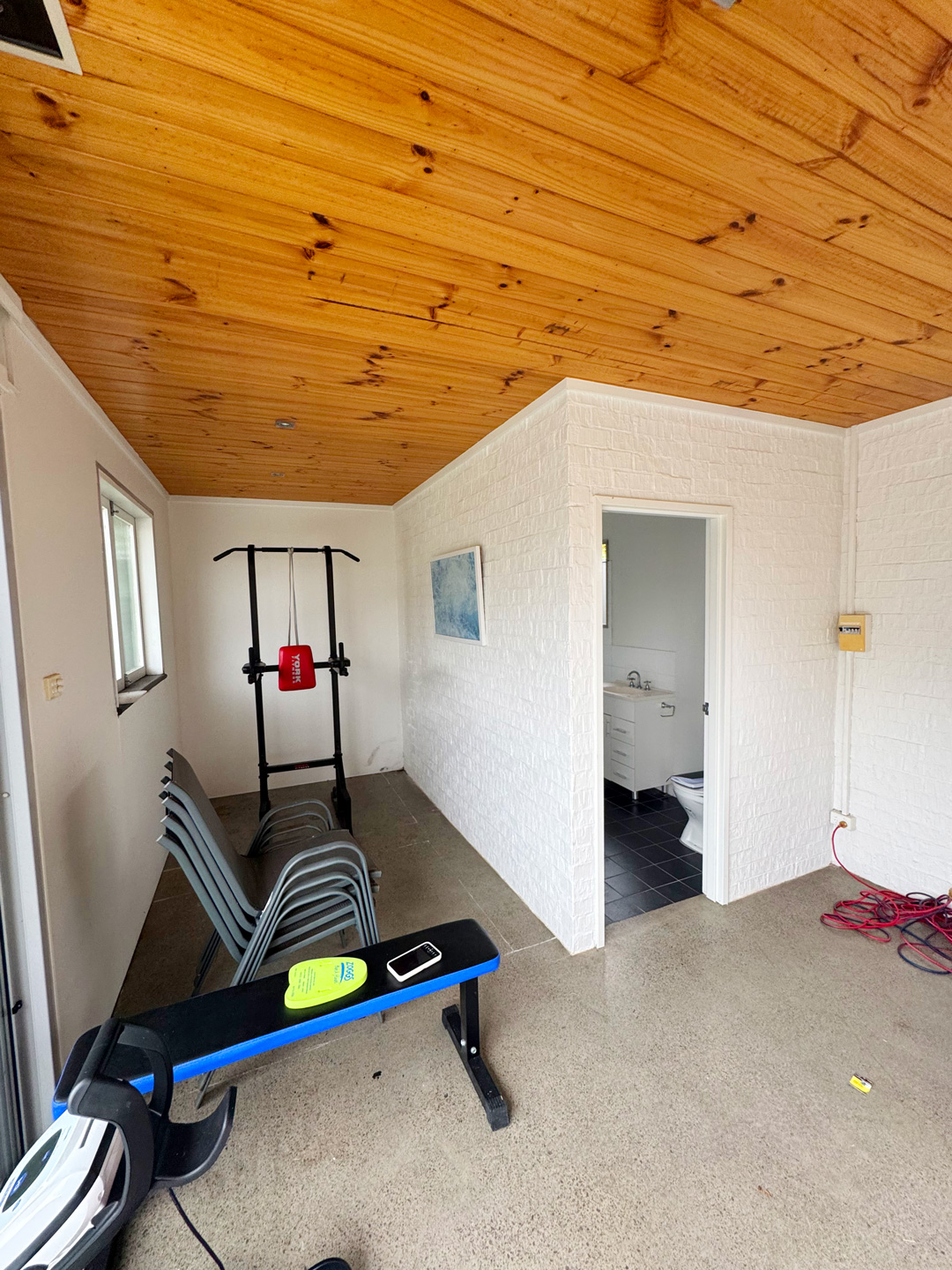 Indoor gym space featuring a pine wood ceiling, white brick walls, and gym equipment including a weight bench and power tower, with an open doorway leading to a bathroom.