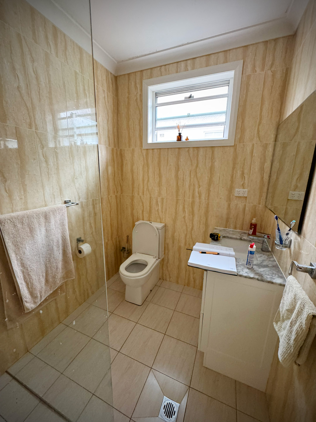 A bathroom featuring tan, vertical-grain wall tiles and a walk-in shower with a glass partition on the left.