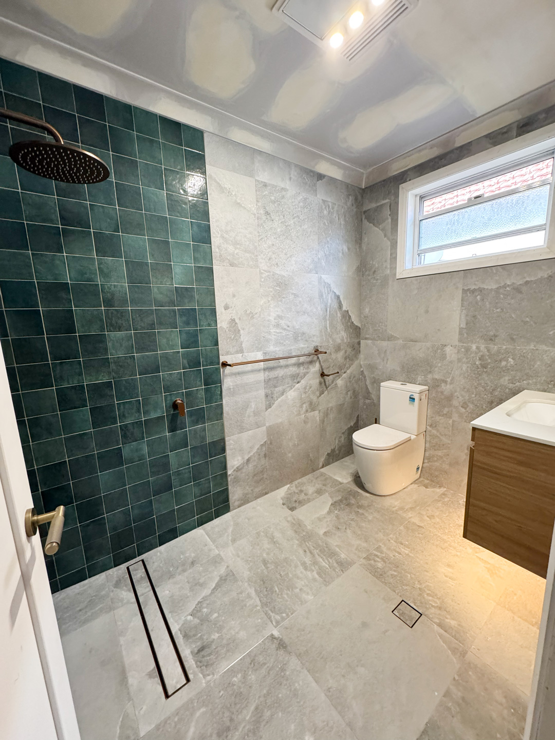 Modern bathroom under renovation featuring a teal square-tiled walk-in shower accent wall paired with large-format grey stone floor and wall tiles