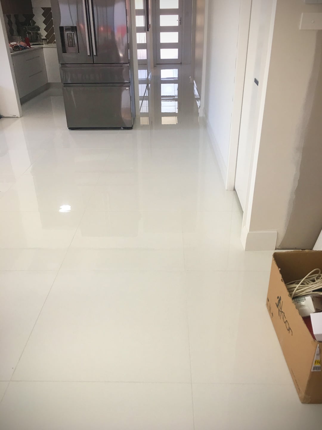 A room with polished, white floor tiles that reflect the overhead lighting and a dark grey refrigerator.