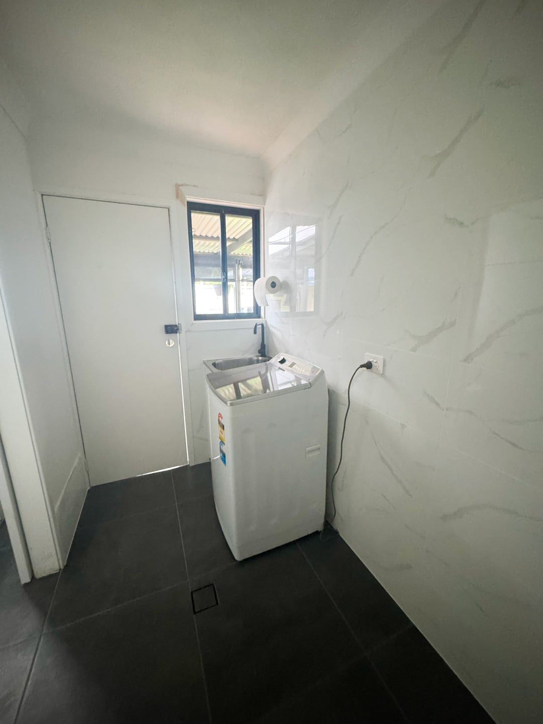 A minimalist laundry room featuring white marbled walls, dark grey floor tiles, a top-loading washing machine, and a small sink under a window.