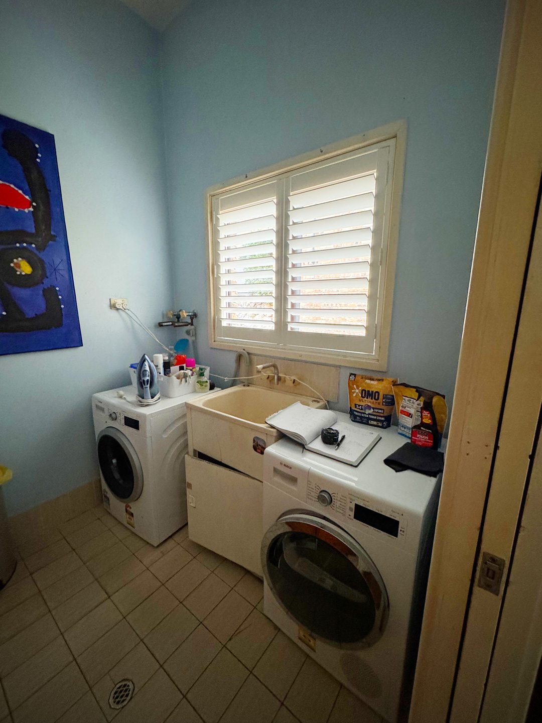 A laundry room with blue walls and tiled floors containing a utility sink between a front-loading washing machine and a dryer, situated under a window with white shutters.