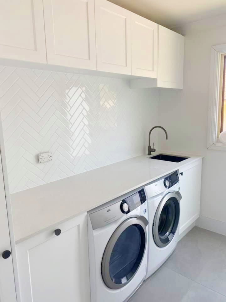 A bright, minimalist all-white laundry room featuring a glossy herringbone tile backsplash, white shaker cabinets.