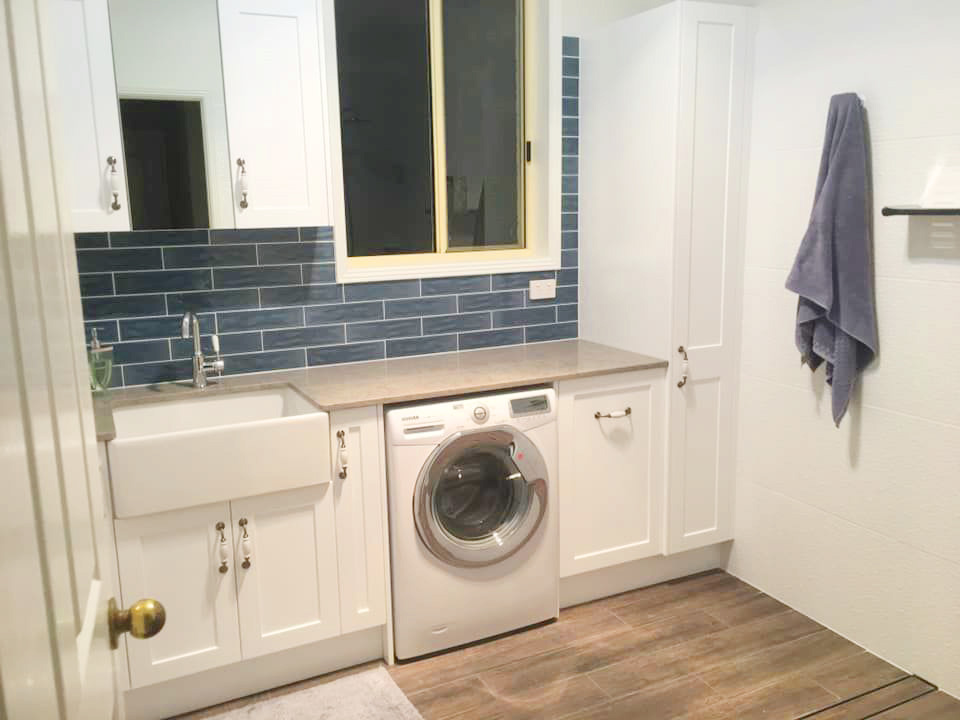 A clean laundry room featuring white shaker-style cabinetry with gold handles, a deep apron-front sink, and wood-look floor tiles.