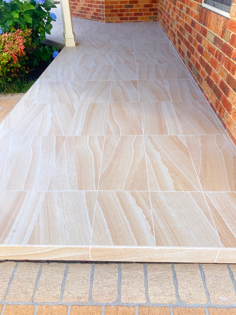 An outdoor porch or walkway featuring large, rectangular tiles with a natural sand-coloured vein pattern.