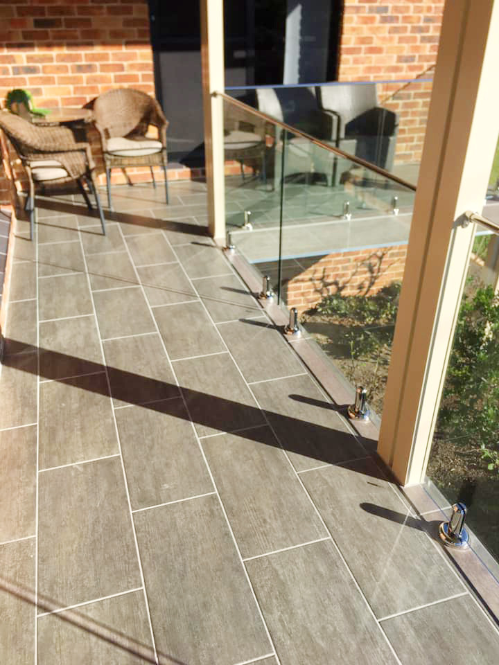 A sunny balcony with grey rectangular tiles laid in a running bond pattern, featuring a glass railing with silver spigots.