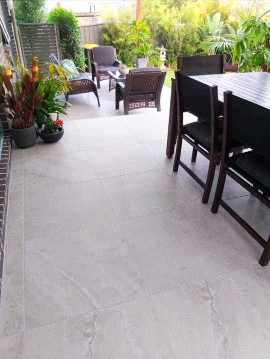 A spacious outdoor patio featuring large, light grey stone-textured tiles, furnished with a dark wood dining set and a comfortable seating area