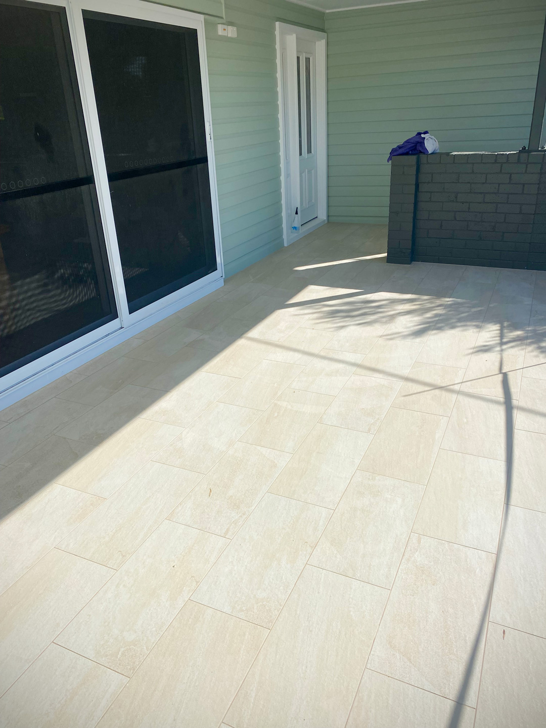 An outdoor patio featuring light-coloured, rectangular tiles laid in a running bond pattern alongside a light green house wall with a white sliding door.