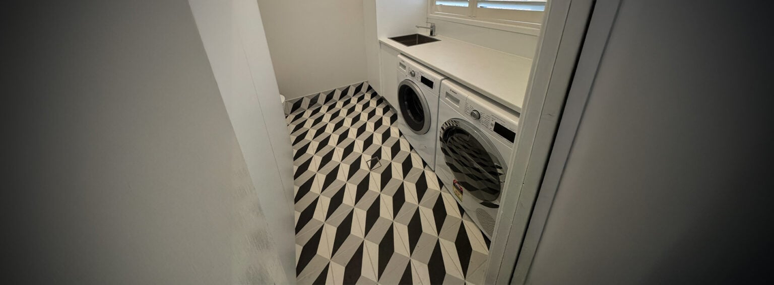 A high-angle, wide shot of a modern laundry room featuring black, white, and grey geometric patterned floor tiles.