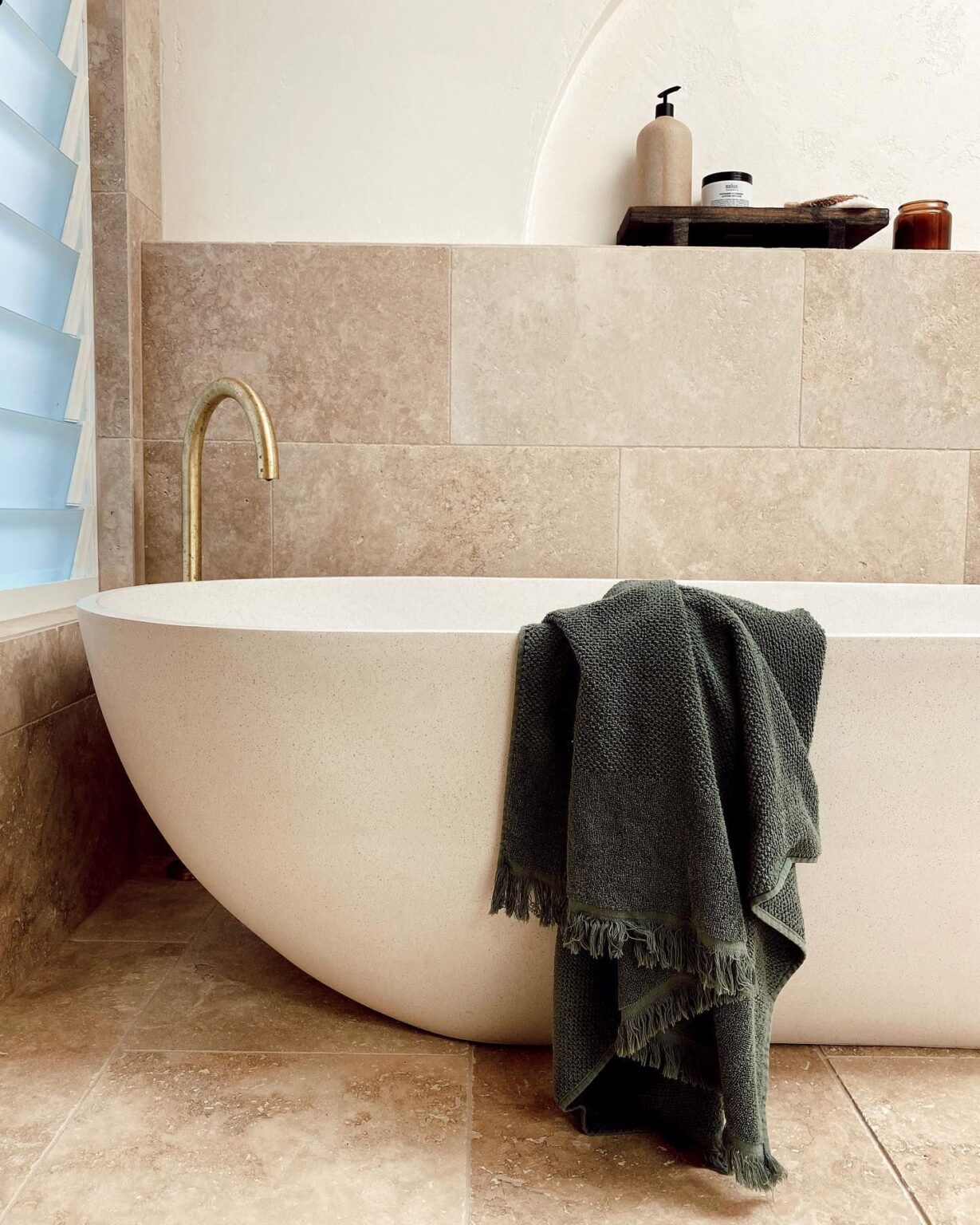 A modern white freestanding bathtub sits against a beige travertine tile wall and floor.