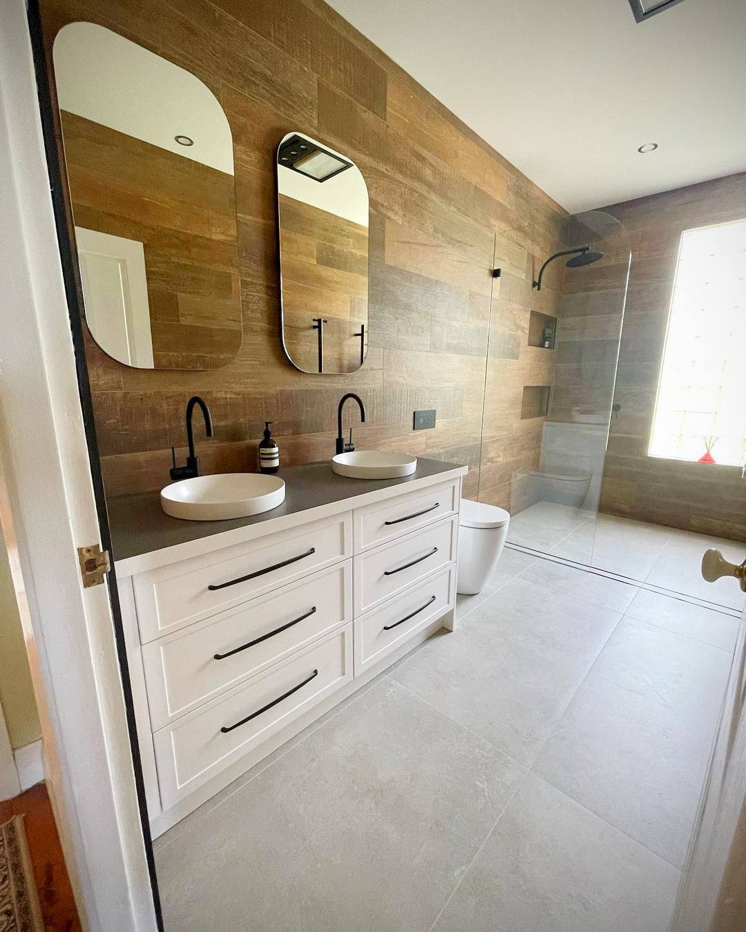 A brightly lit, contemporary bathroom showing a white vanity with two vessel sinks and black arched mirrors against wood-plank walls.