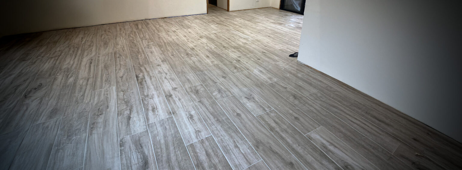 A wide, low-angle shot of a room featuring light grey wood-look floor tiles laid in a staggered plank pattern.