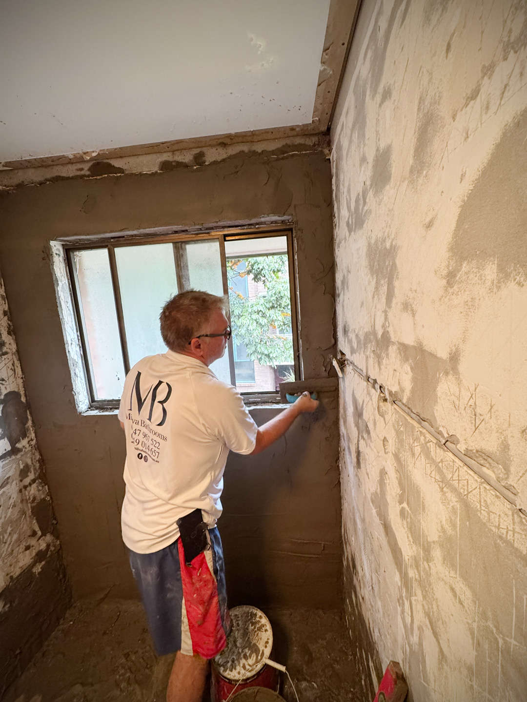 A worker in a white t-shirt applying a dark grey cement render or plaster to a bathroom wall around a small window using a hand trowel.