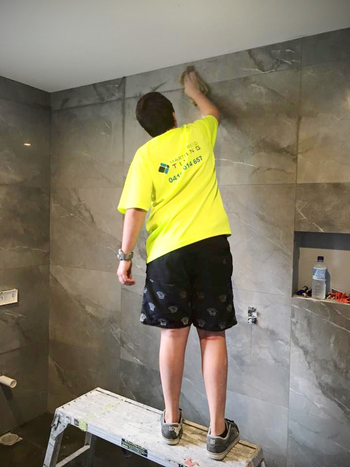 A person wearing a Martin Reid Tiling high-visibility yellow shirt stands on a small step ladder, wiping down large, grey marble-patterned wall tiles.