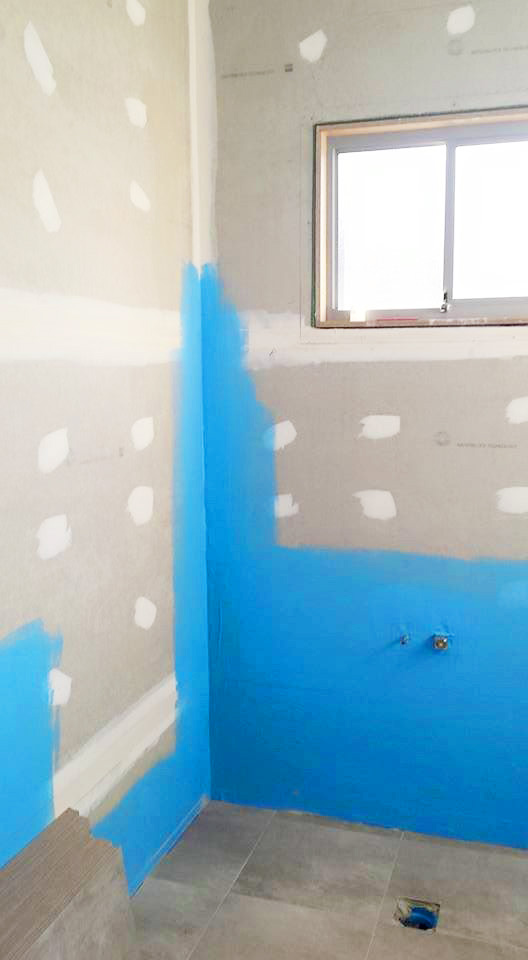 An interior corner of a bathroom under construction, featuring grey wall panels with white joint compound and a bright blue waterproofing membrane