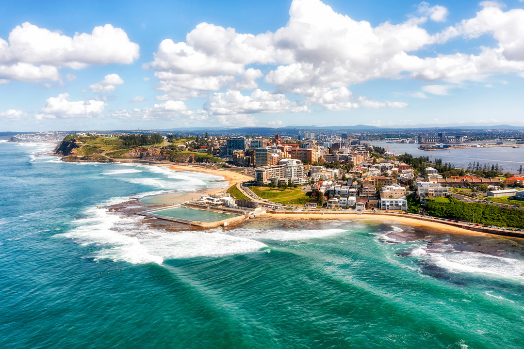 An aerial view of a vibrant coastal city featuring turquoise ocean waves crashing against a rocky shoreline and an ocean bath.